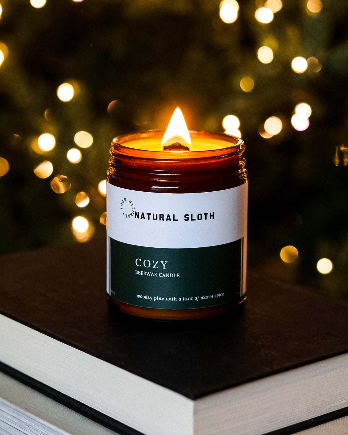 A lit jar candle labeled Natural Sloth Cozy  Beeswax Candle sits on top of stacked books, with a blurred Christmas tree of warm lights in the background.