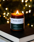 A lit jar candle labeled Natural Sloth Cozy  Beeswax Candle sits on top of stacked books, with a blurred Christmas tree of warm lights in the background.