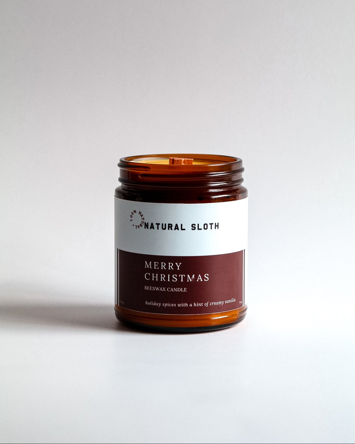 An amber glass jar Beeswax candle with a white and dark red label reading 'Natural Sloth Merry Christmas', photographed on a clean light gray background.