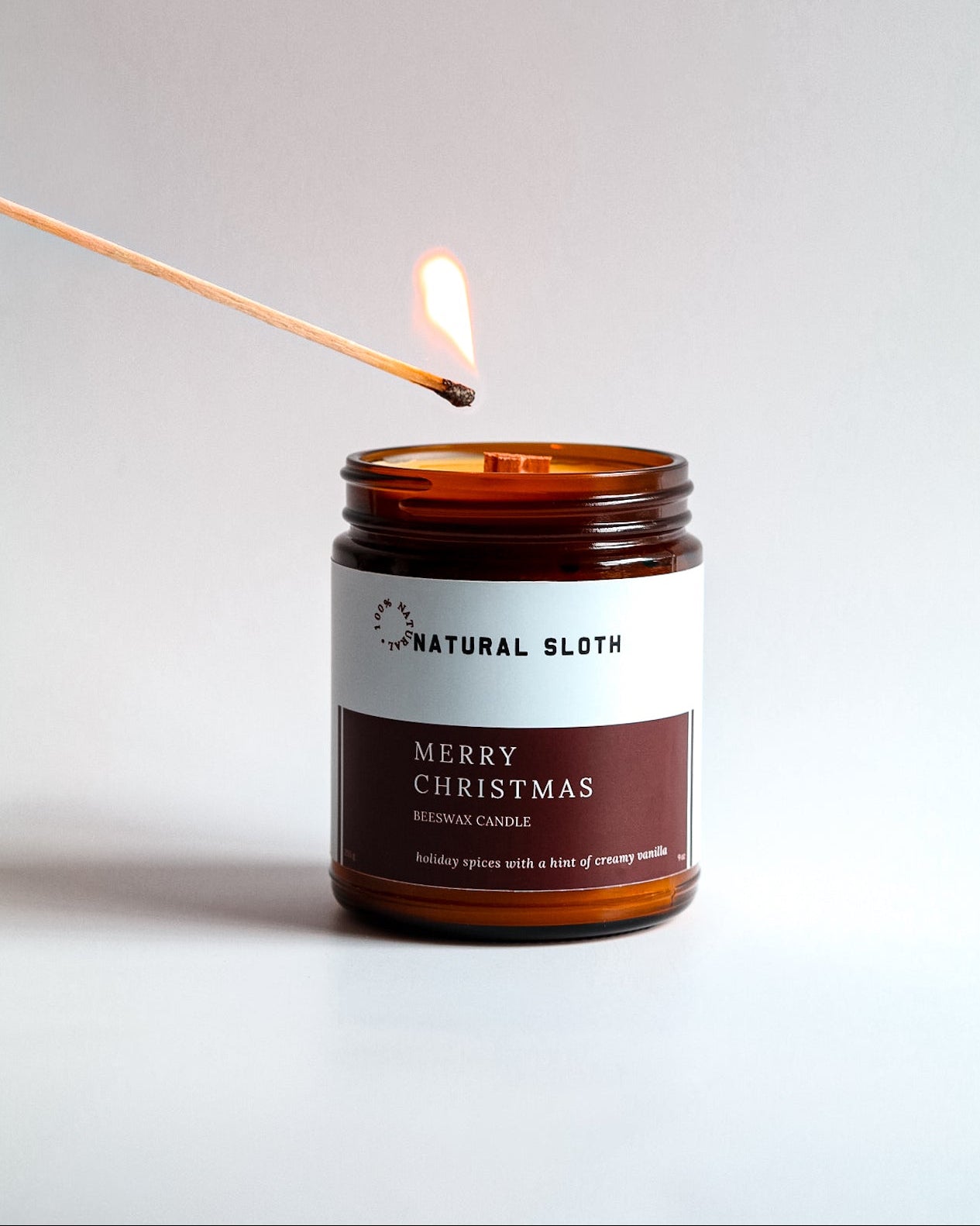 An amber glass jar Beeswax candle labeled Natural Sloth Merry Christmas, with a lit match held above it against a plain light background.