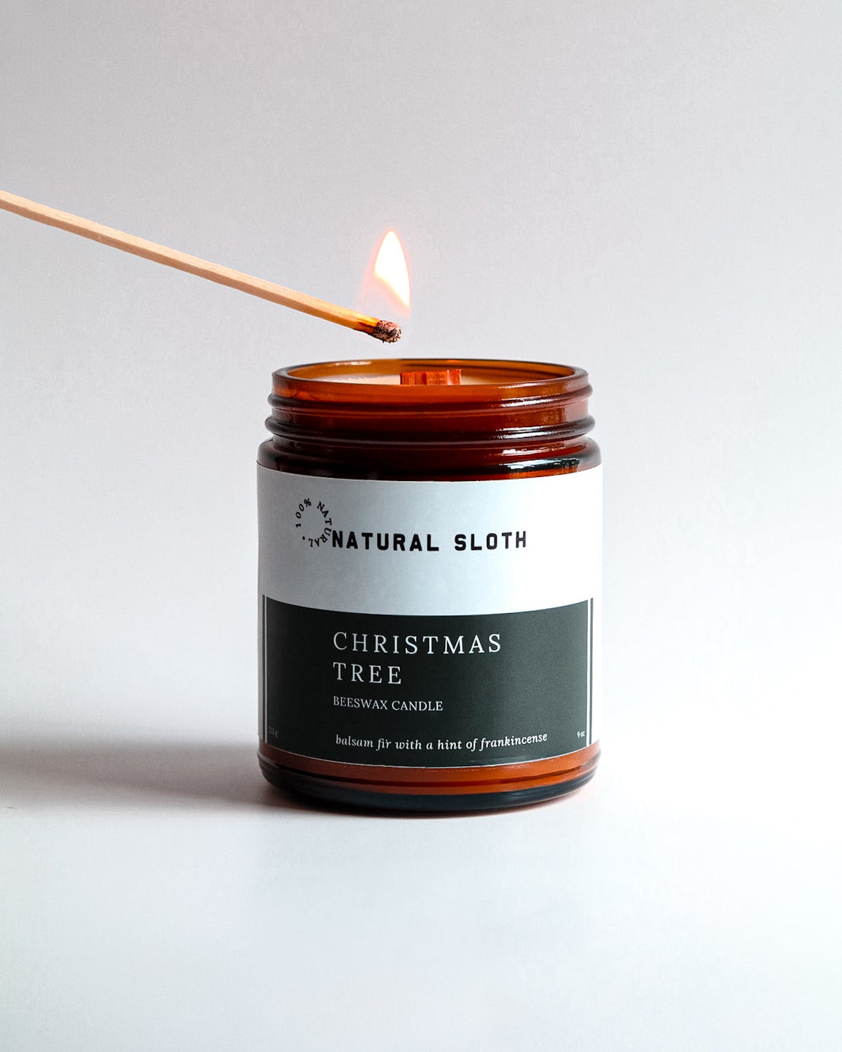 An amber glass jar candle labeled 'Natural Sloth Christmas Tree Beeswax Candle' is lit with a wooden match against a clean white background.