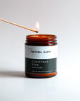 An amber glass jar candle labeled 'Natural Sloth Christmas Tree Beeswax Candle' is lit with a wooden match against a clean white background.