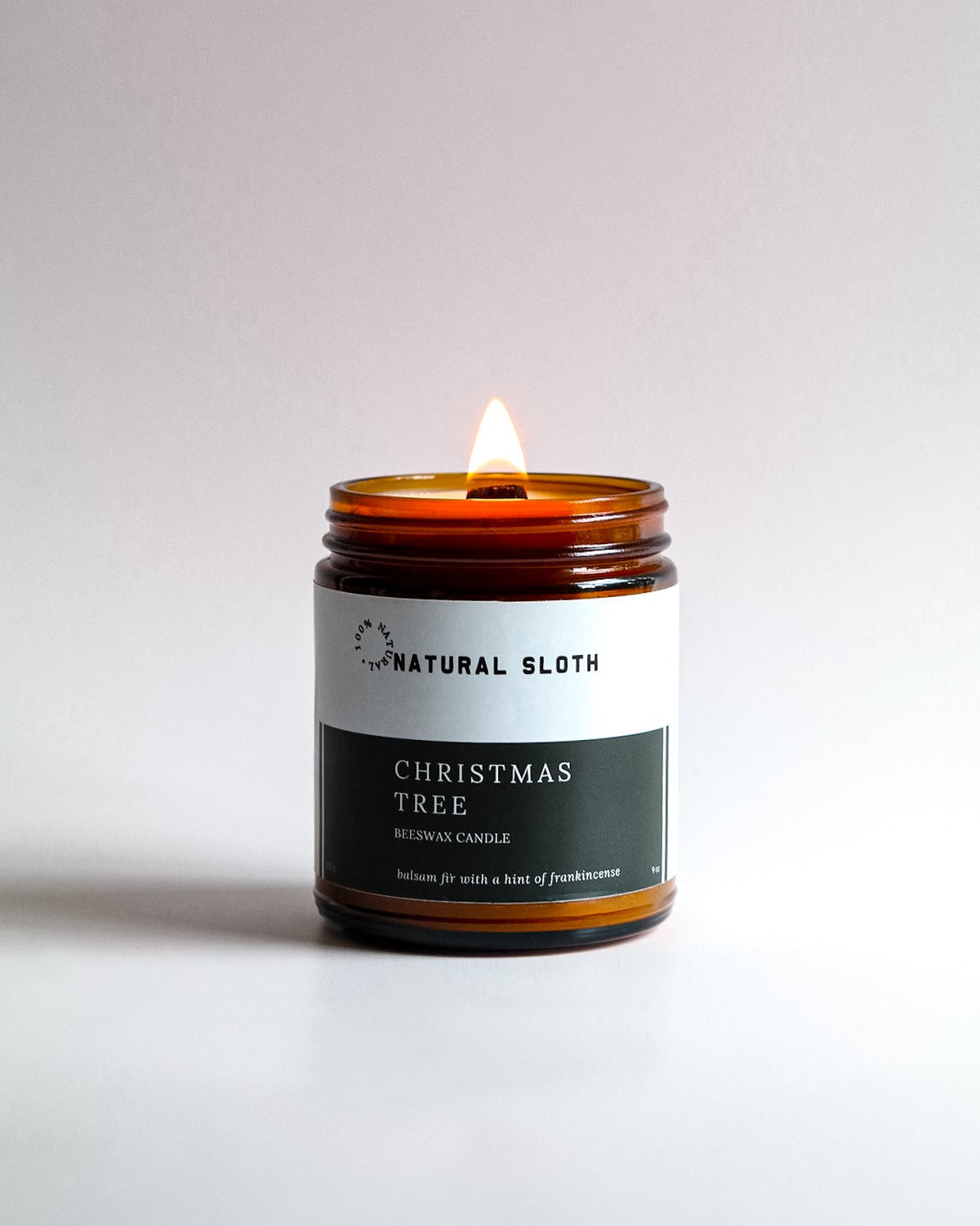 A lit amber glass jar candle with a white and dark green label reading 'Natural Sloth Christmas Tree Beeswax Candle', set against a clean light gray background.