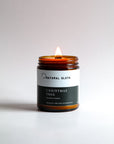 A lit amber glass jar candle with a white and dark green label reading 'Natural Sloth Christmas Tree Beeswax Candle', set against a clean light gray background.