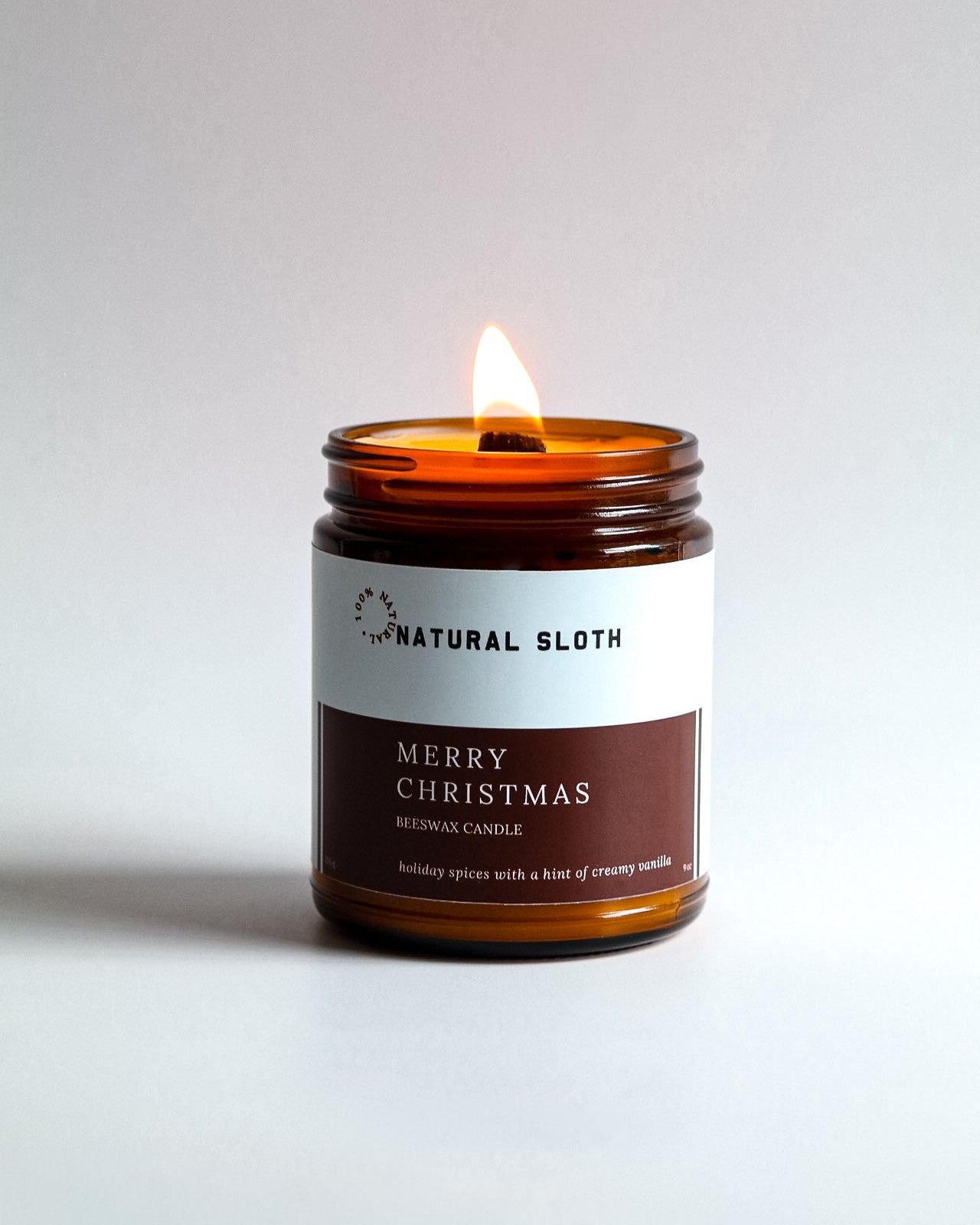 A lit Natural Sloth 'Merry Christmas' scented Beeswax candle in an amber glass jar, set against a clean, light gray background.