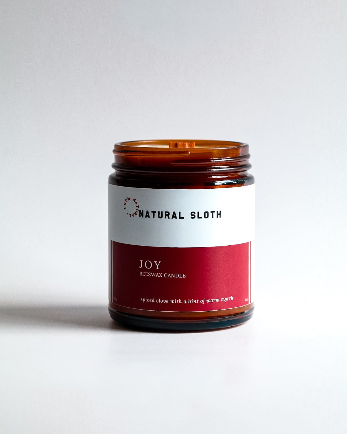 A Natural Sloth 'Joy' Beeswax candle in an amber glass jar, featuring a white and red label, set against a clean light gray background.