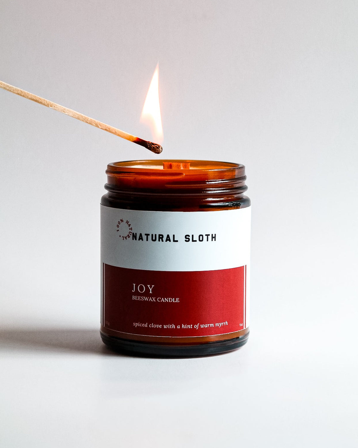 An amber glass jar candle labeled 'Natural Sloth Joy Beeswax Candle' with a lit match hovering above it on a clean white background.