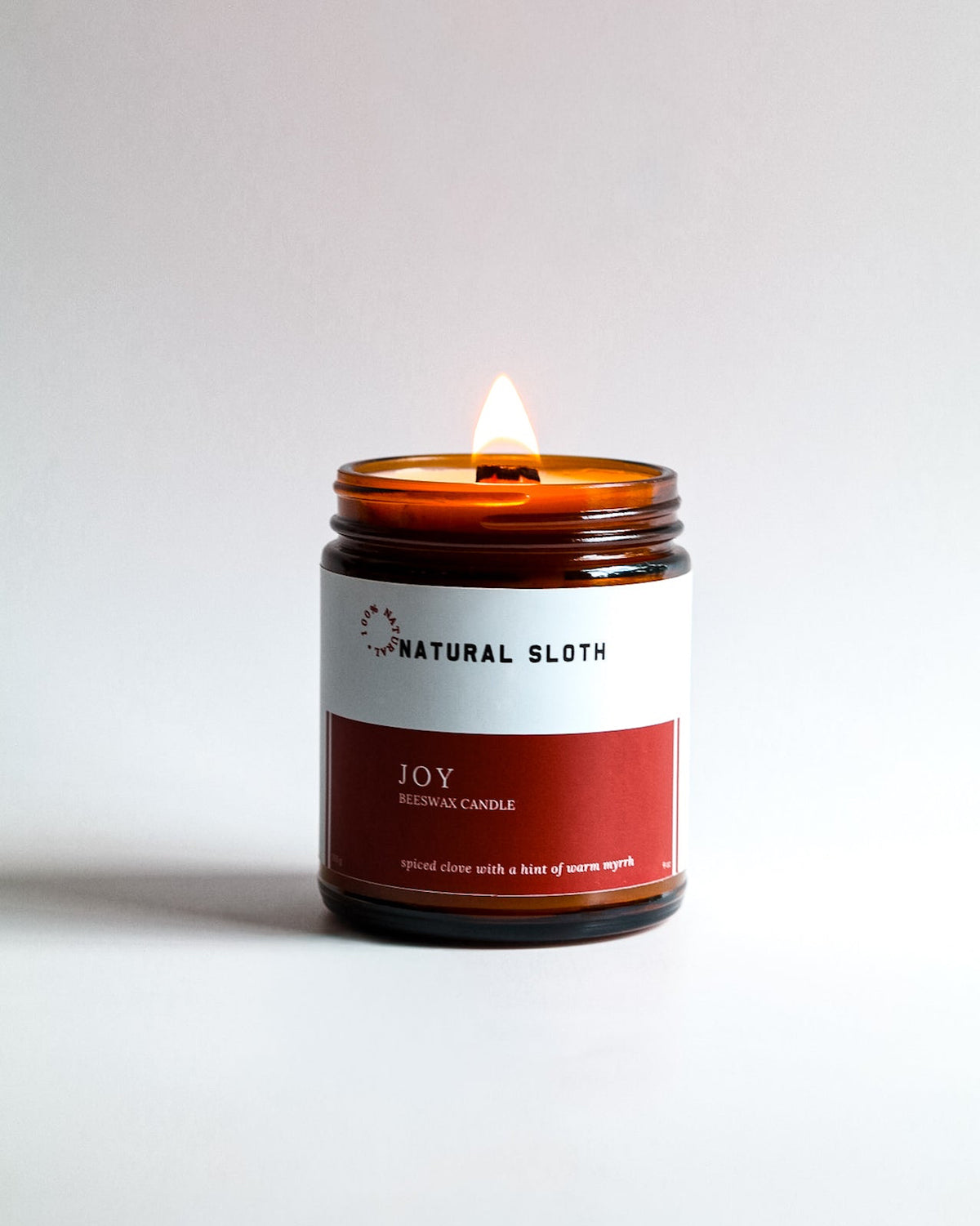 A lit amber glass jar Beeswax candle with a white and red label reading 'Natural Sloth Joy Scented Candle' on a clean white background.