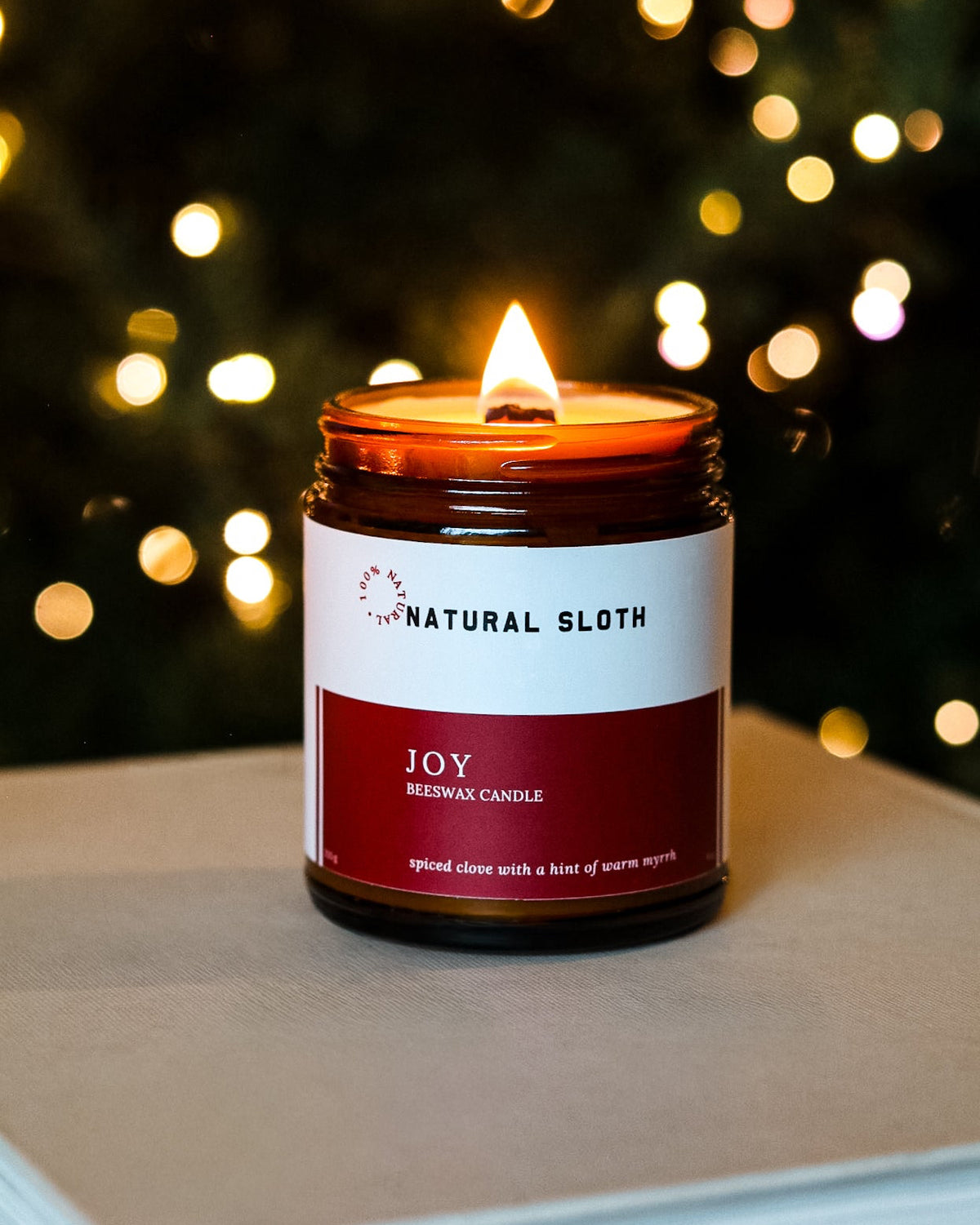 A lit Natural Sloth 'Joy' Beeswax Candle in an amber glass jar, resting on a light-colored book with a festive, blurred Christmas tree background of warm bokeh lights.