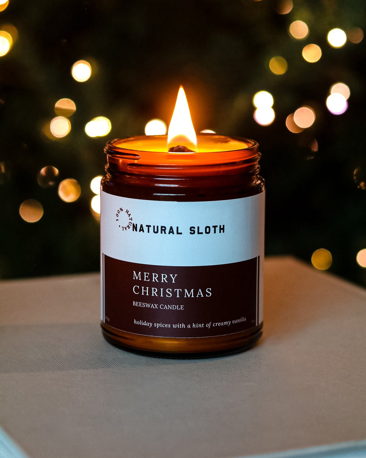 A lit Natural Sloth Mery Christmas candle in an amber glass jar sits on a neutral surface with a blurred Christmas tree and warm bokeh lights in the background.