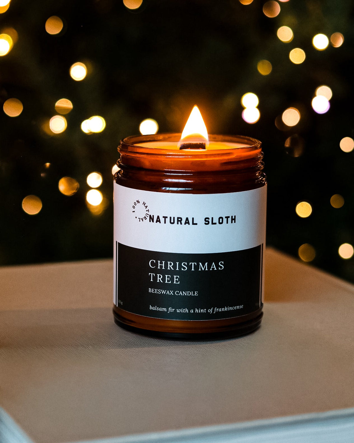 A lit amber glass candle jar labeled Natural Sloth Christmas Tree Beeswax Candle, placed on a neutral surface with a blurred festive Christmas Tree background of warm lights.