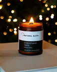 A lit amber glass candle jar labeled Natural Sloth Christmas Tree Beeswax Candle, placed on a neutral surface with a blurred festive Christmas Tree background of warm lights.