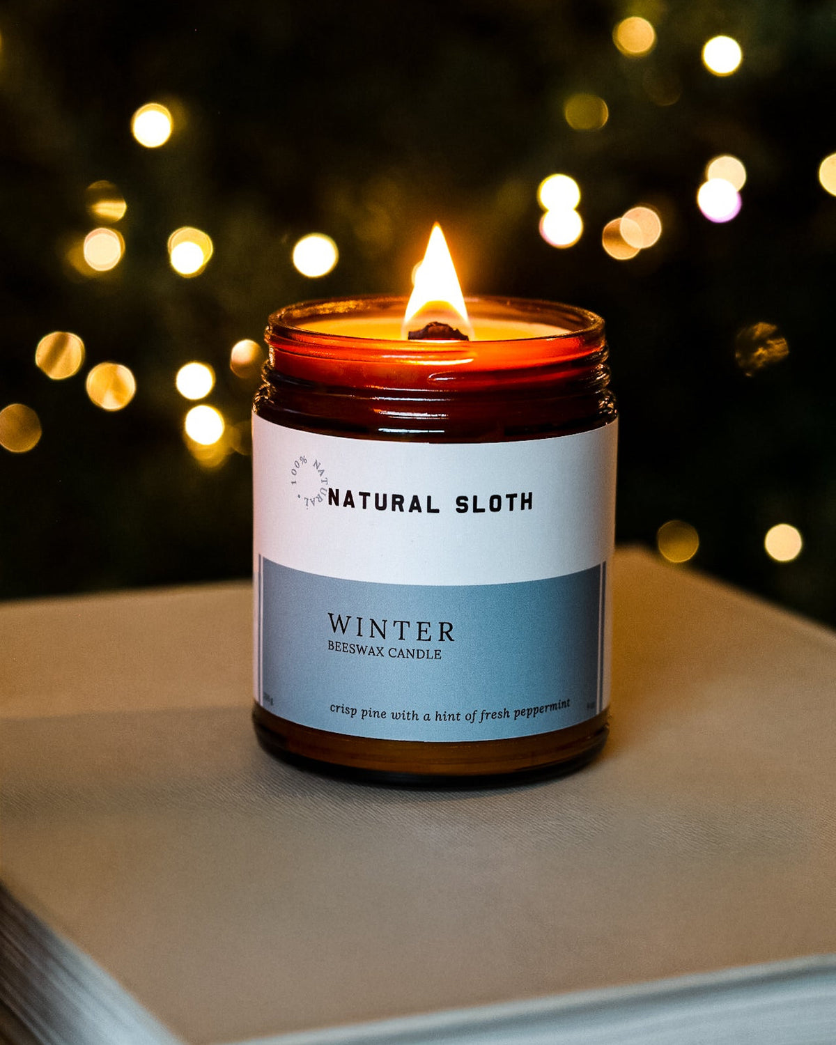 A lit Natural Sloth Winter Beeswax Candle in an amber glass jar, resting on a neutral-book with a festive, blurred Christmas tree and warm bokeh lights in the background.